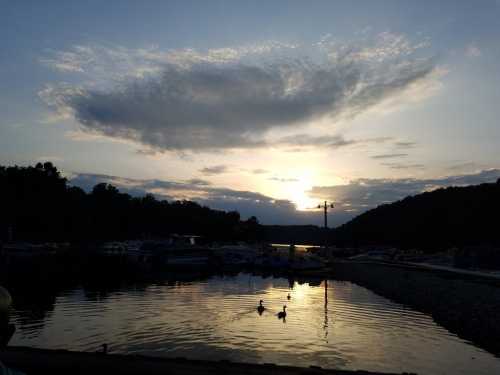 Sunset over a calm lake with boats and ducks silhouetted against the colorful sky and clouds.