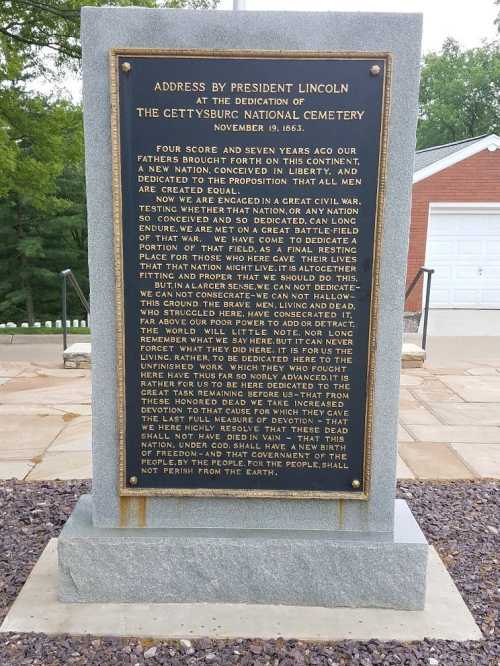 A stone monument with an inscription of President Lincoln's address at Gettysburg National Cemetery, surrounded by greenery.