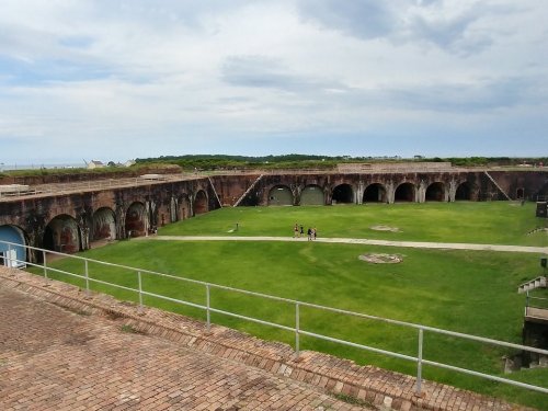 A historic fort with arched openings, surrounded by green grass and a cloudy sky, with a few people walking inside.