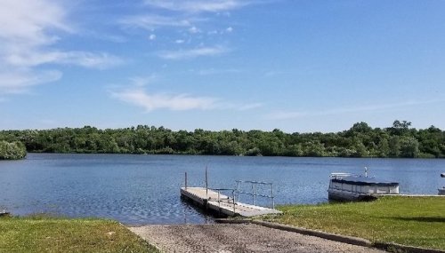 A serene lake view with a wooden dock, surrounded by lush greenery and a clear blue sky.