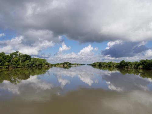 A serene river scene with lush green banks, reflecting clouds and trees in calm water under a partly cloudy sky.