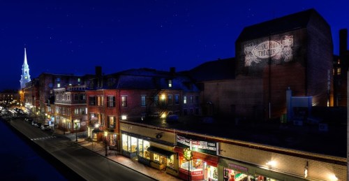 A nighttime view of a historic street with shops, illuminated buildings, and a church steeple in the background.
