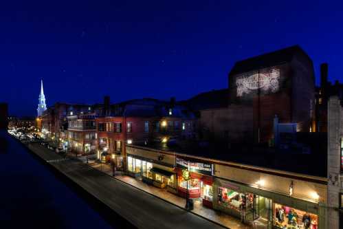 Nighttime view of a charming street with shops, illuminated buildings, and a church steeple against a starry sky.