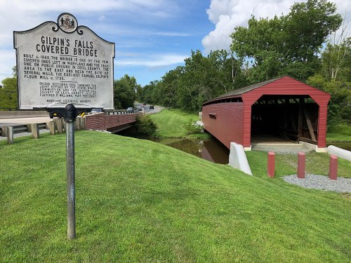 A historic sign next to Gilpin's Falls Covered Bridge, with a grassy area and a road in the background.
