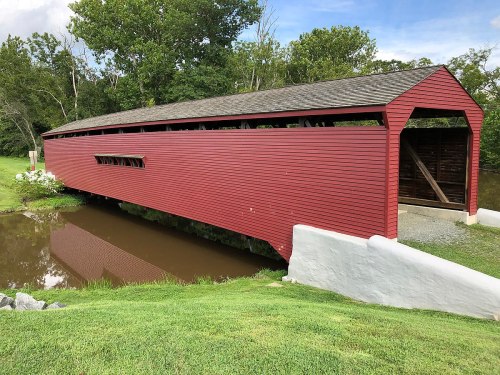 A red covered bridge spans a calm pond, surrounded by green grass and trees under a partly cloudy sky.