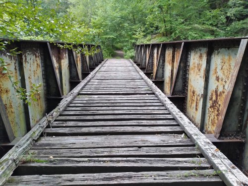 A weathered wooden bridge with rusty metal supports, surrounded by lush green trees and foliage.