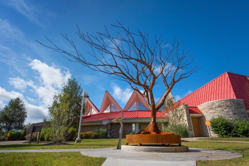 A bare tree sculpture stands in front of a modern building with a red roof, under a blue sky with scattered clouds.