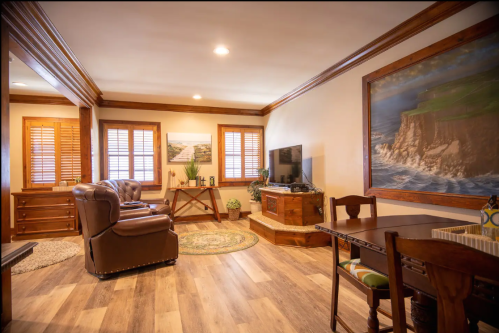 Cozy living room with a brown armchair, wooden furniture, a TV, and a scenic wall painting. Warm lighting and wooden accents.