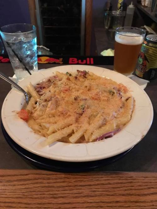 A plate of creamy pasta topped with cheese, accompanied by a glass of water and two beers on a bar counter.