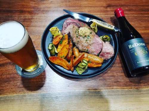 A plate of roasted meat with vegetables, a glass of beer, and a bottle of ReDone beer on a wooden table.