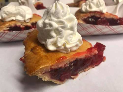 A slice of cherry pie topped with whipped cream, surrounded by more pie slices in the background.