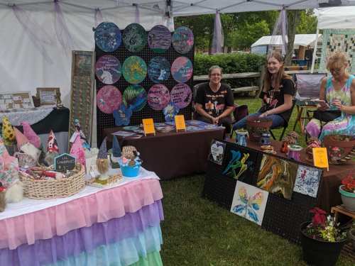 A craft fair booth with colorful artwork, handmade items, and three women engaging with visitors under a tent.