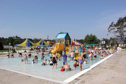 A busy outdoor pool scene with families and children playing, colorful slides, and sun umbrellas in the background.