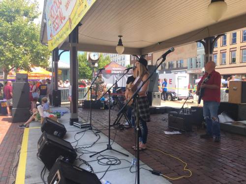 A band performs on stage at a festival, with a crowd enjoying the event in the background.