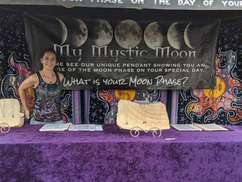 A woman stands behind a table displaying moon phase pendants, with a colorful banner reading "My Mystic Moon."