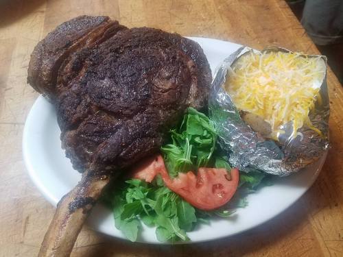 A large, grilled ribeye steak on a plate with fresh greens, tomato slices, and a baked potato topped with cheese.