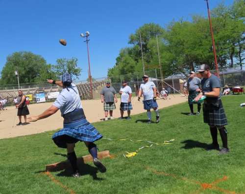A group of men in kilts compete in a field event, throwing a heavy stone on a sunny day.