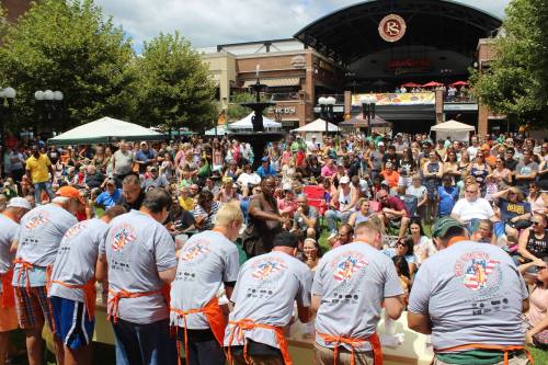 A crowd watches as contestants compete in a food-eating contest outdoors, with tents and trees in the background.