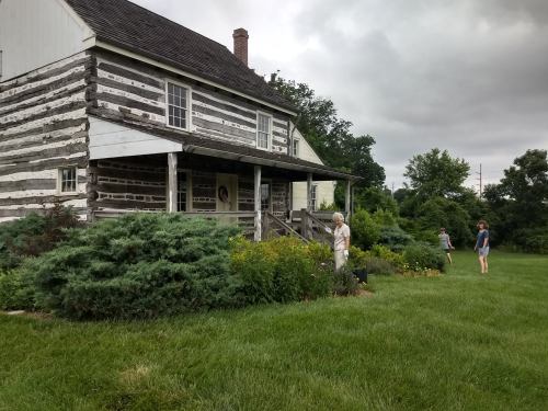 A rustic log cabin surrounded by greenery, with two people tending to the garden and one walking in the background.