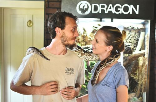 A man and woman share a close moment, with a snake draped around the man's neck in a cozy indoor setting.