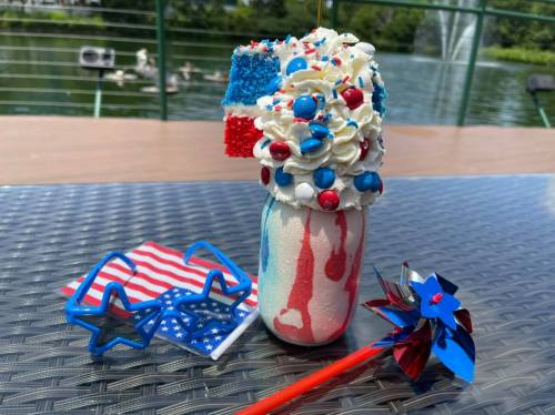 A festive milkshake topped with whipped cream, red, white, and blue decorations, alongside patriotic sunglasses and a pinwheel.