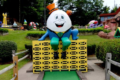 A large cartoonish egg character in a top hat sits on a colorful fence at a mini-golf course surrounded by whimsical scenery.