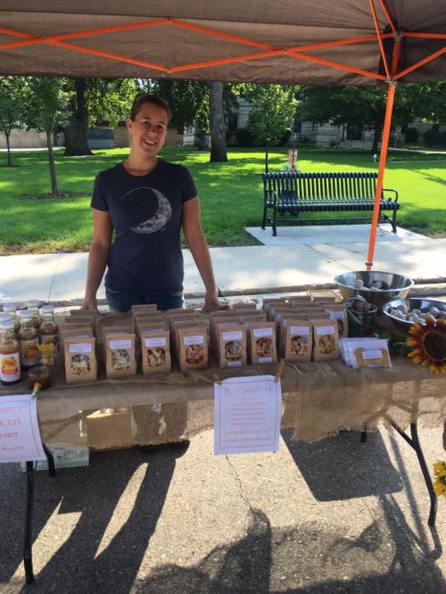 A smiling vendor stands behind a table of packaged goods at an outdoor market, with a park in the background.