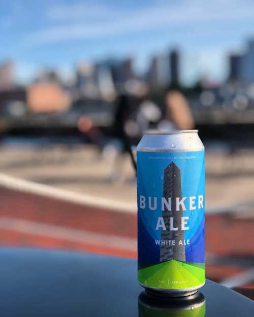 A can of Bunker Ale White Ale sits on a table with a city skyline in the background on a sunny day.