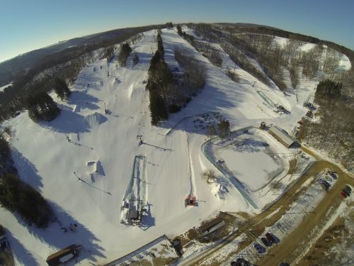 Aerial view of a snowy ski resort with slopes, trees, and a parking area under a clear blue sky.