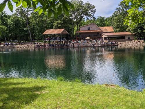 A serene lake scene with a wooden dock and a building, surrounded by lush greenery and people enjoying the outdoors.