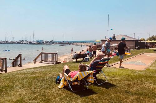 People relax on beach chairs by the water, with boats and a marina in the background on a sunny day.