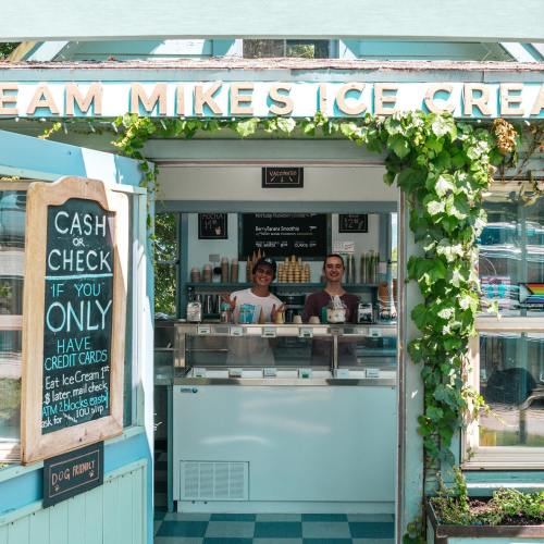 A charming ice cream shop with two staff members behind the counter, surrounded by greenery and a sign for payment options.