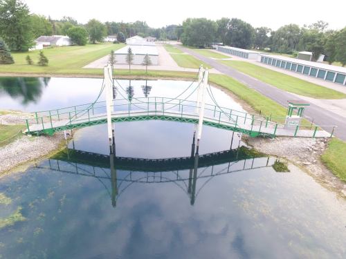 A green pedestrian bridge spans a calm pond, reflecting in the water, with storage units and trees in the background.