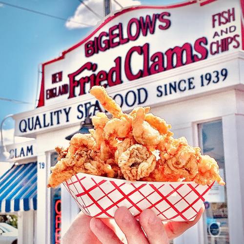 A hand holds a basket of fried clams in front of a seafood restaurant sign that reads "Bigelow's Fried Clams."