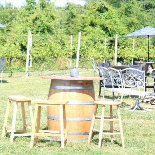 A wooden barrel table surrounded by three stools in a vineyard setting with green foliage in the background.