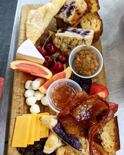A wooden platter with assorted fruits, cheeses, baked goods, and a small bowl of sauce.