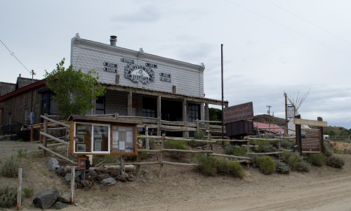 A rustic building with a wooden fence, signs, and a dirt path, set against a cloudy sky in a small town.