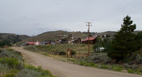 A dirt road leads to a small, rustic town with wooden buildings and hills in the background under a cloudy sky.