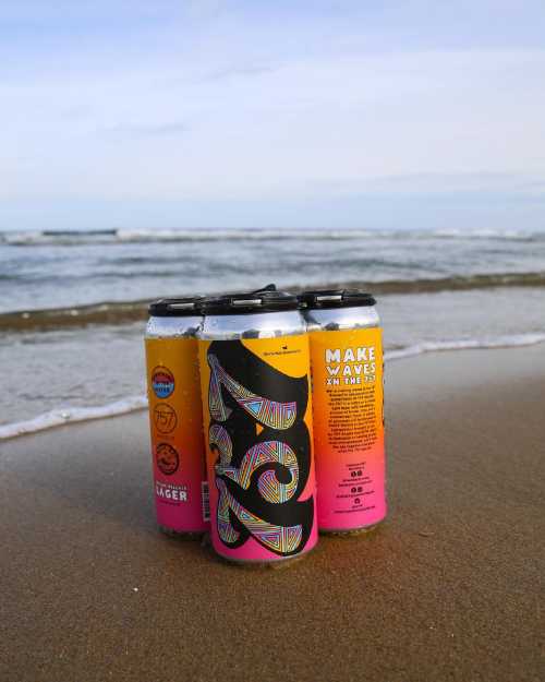 Four colorful cans of beer on a sandy beach with ocean waves in the background under a clear sky.