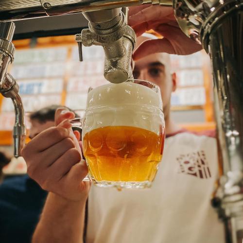 A person pours a frothy beer into a glass from a tap, with a colorful background of beer labels.