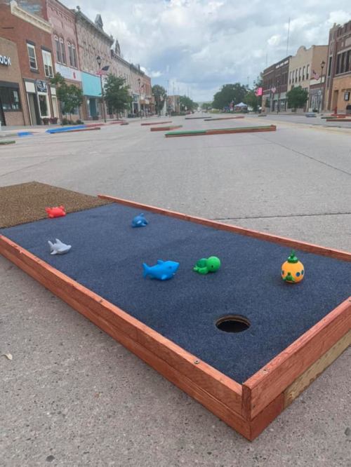Colorful mini-golf course setup on a street, featuring toy sea creatures and a hole on a blue carpeted surface.