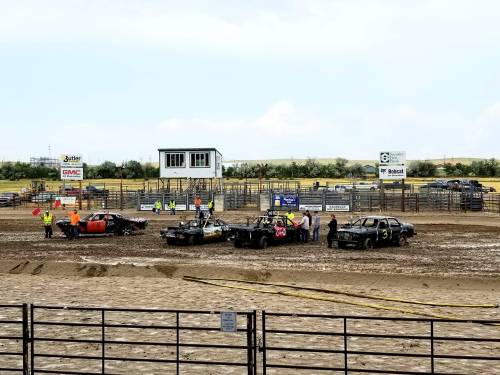 A muddy demolition derby scene with several damaged cars and spectators in safety vests at a racetrack.