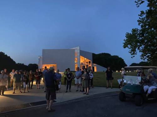 A crowd gathers outside a modern building at dusk, with some people seated in a golf cart nearby.