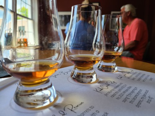Three whiskey glasses filled with amber liquid on a table, with blurred patrons in the background.