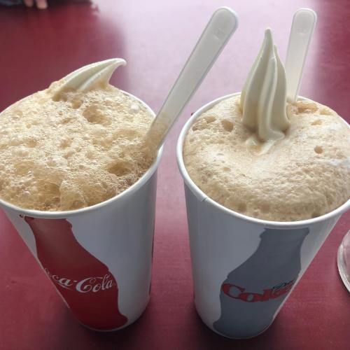 Two cups of frothy root beer floats topped with soft serve ice cream, featuring classic Coca-Cola branding.