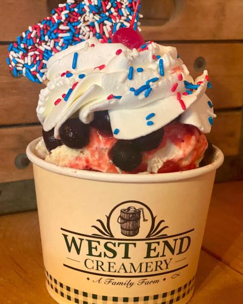 A cup of ice cream topped with whipped cream, sprinkles, cherries, and mixed berries, labeled "West End Creamery."