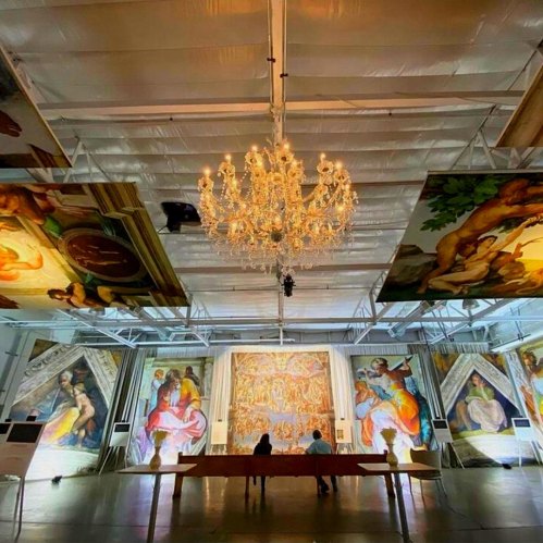 A spacious gallery with large illuminated artworks and a chandelier, featuring two people observing the displays.