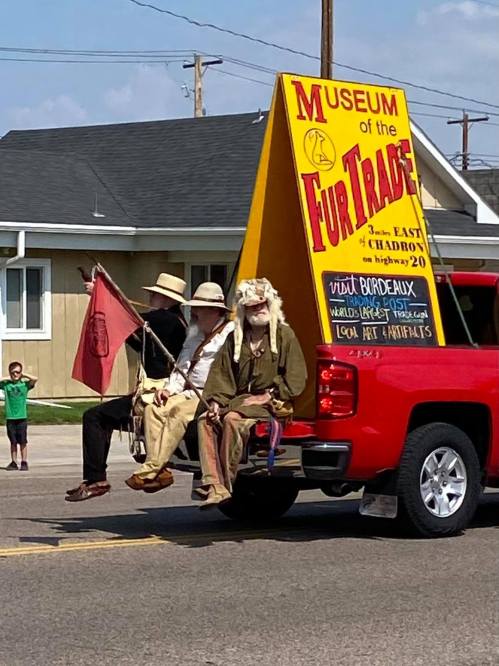 Three people in historical attire sit in the back of a red truck, promoting the Museum of the Fur Trade.