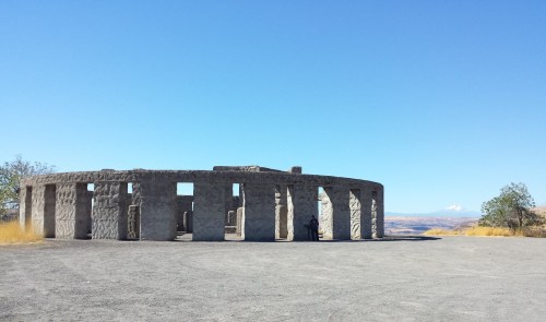 A stone structure resembling Stonehenge against a clear blue sky, surrounded by dry grass and distant mountains.