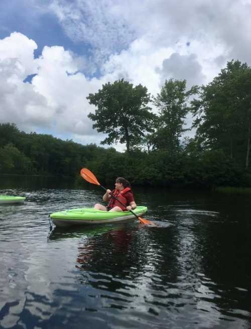 A person in a red life jacket paddles a green kayak on a calm river surrounded by trees and clouds.